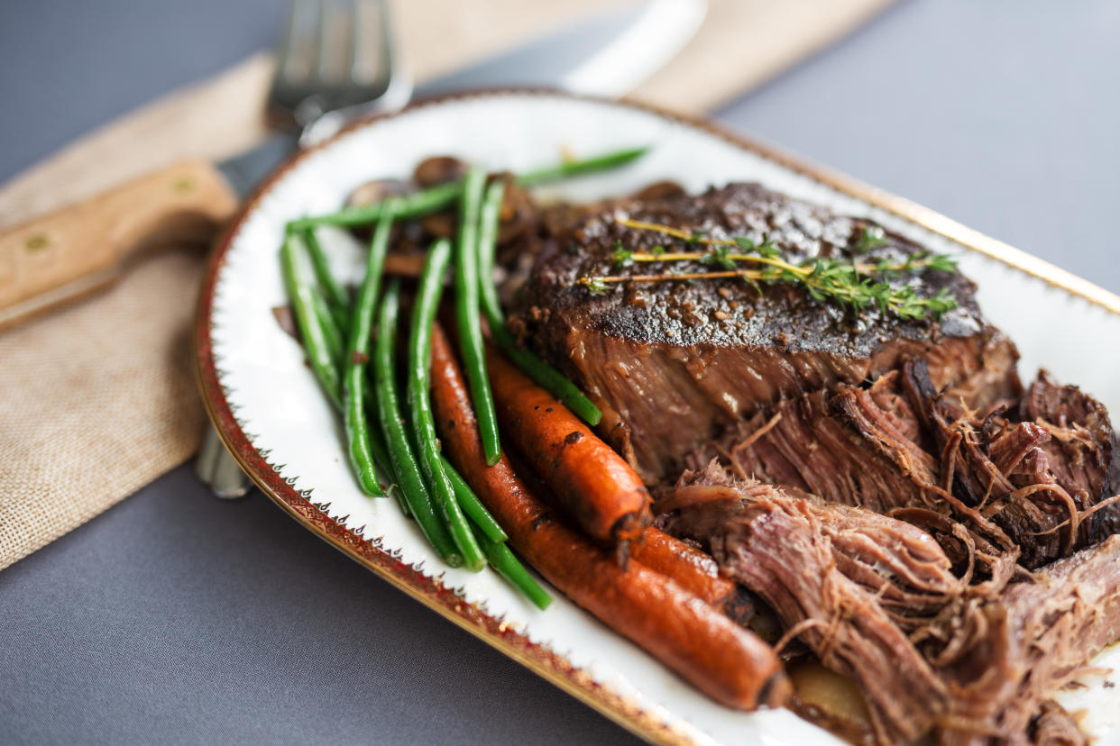 Pot roast being served on a white oval platter with gold rim. Gray tablecloth. Copy space.