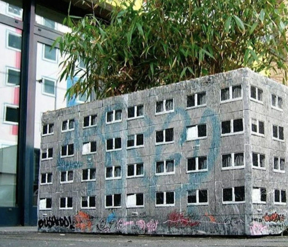 Miniature cinder block-style artwork with graffiti, resembling a building, placed in front of a bush