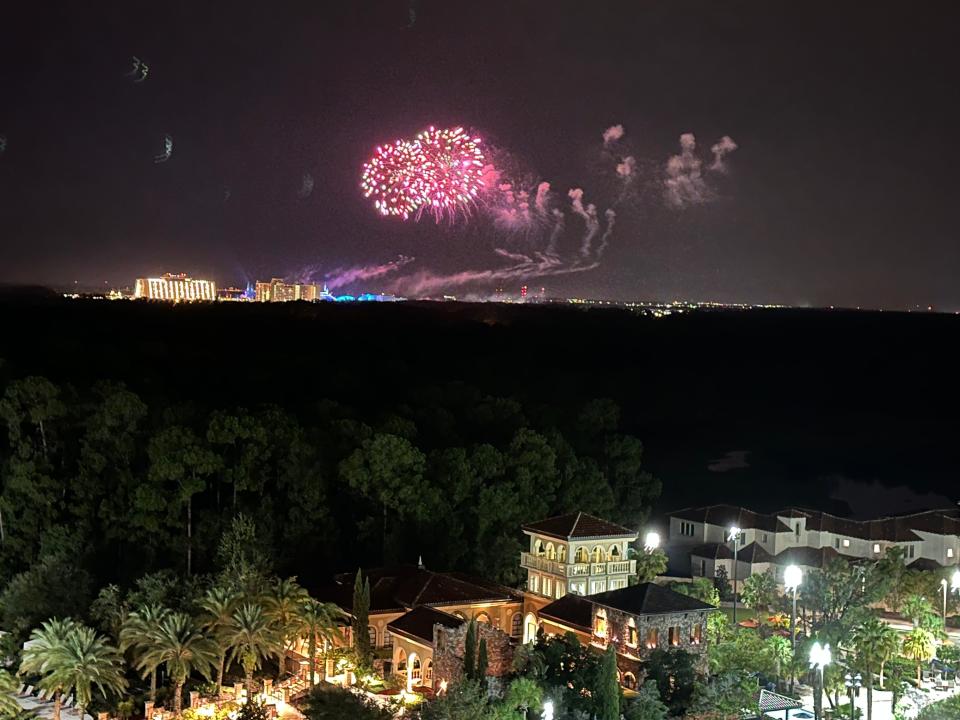 Fireworks in the distance are bright red and gold. In the foreground, the Disney theme park is lit up at night, surrounded by palm trees.