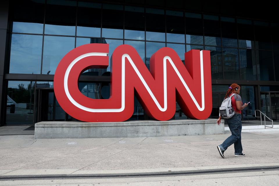 Plastic CNN red logo in front of a building. A woman in jeans and a red t-shirt is walking by on the pavement.