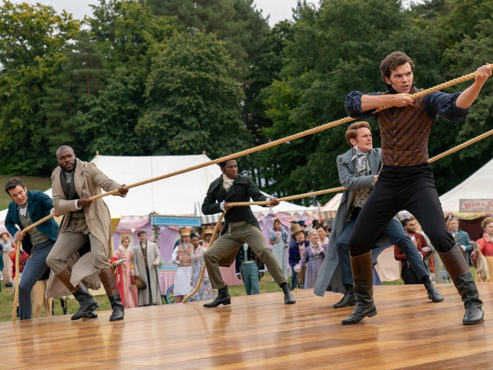 colin bridgerton and other men standing on a wooden platform, pulling at several ropes tethered to a balloon. colorfully dressed onlookers watch from the background