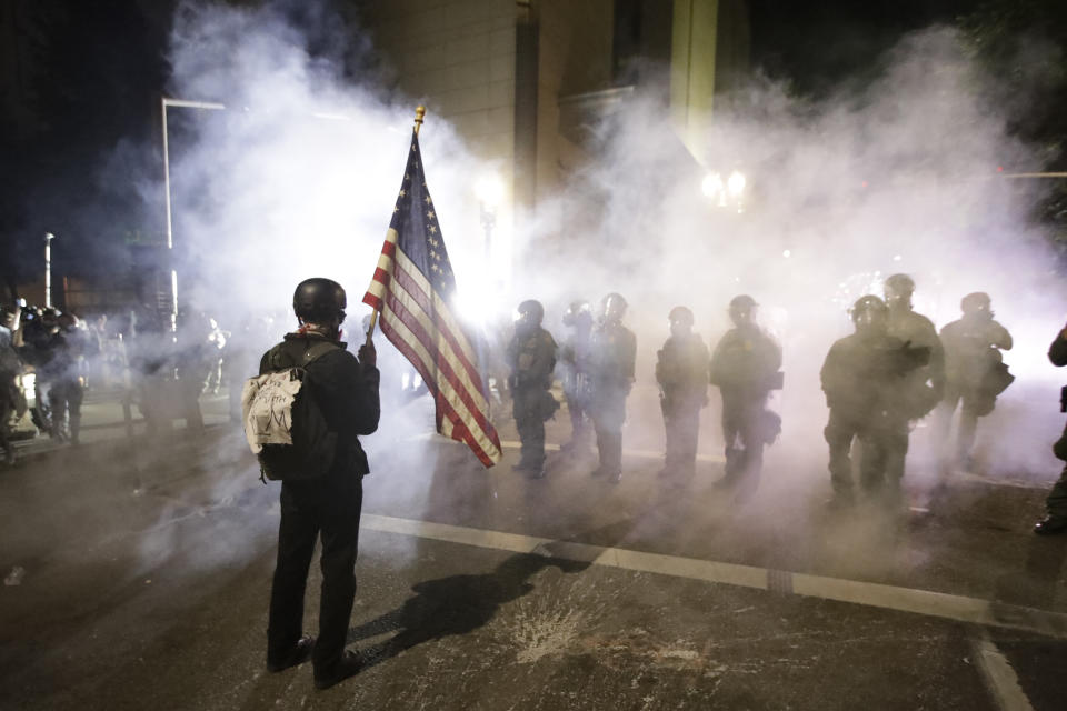 A demonstrator waves a U.S. flags in front of federal agents after tear gas is deployed during a Black Lives Matter protest at the Mark O. Hatfield United States Courthouse Thursday, July 30, 2020, in Portland, Ore. (AP Photo/Marcio Jose Sanchez)