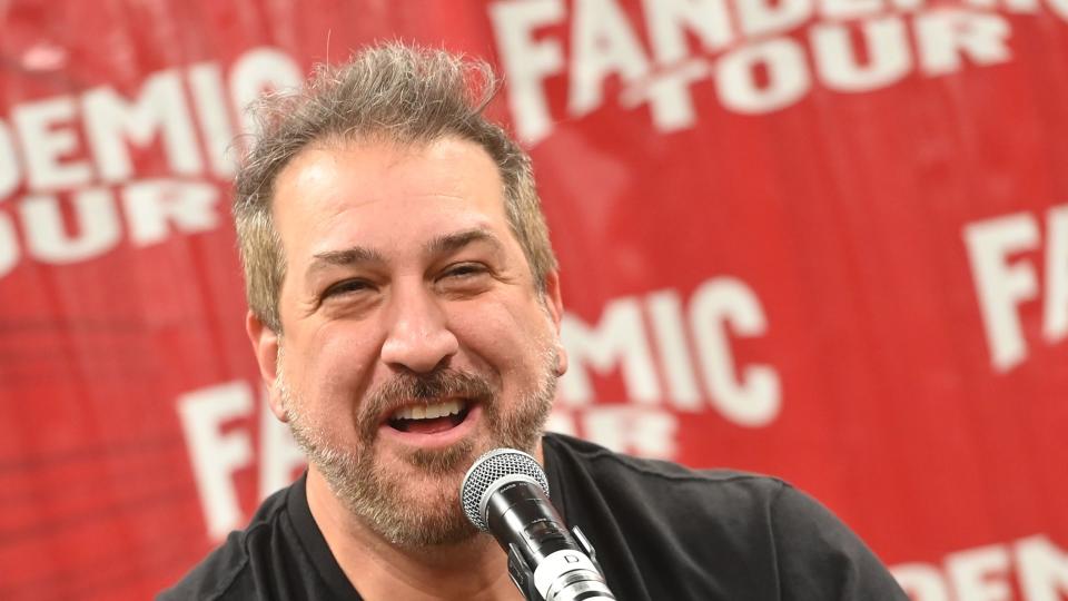 joey fatone smiling as he speaks into a microphone seated at a table and holding a bottle of water