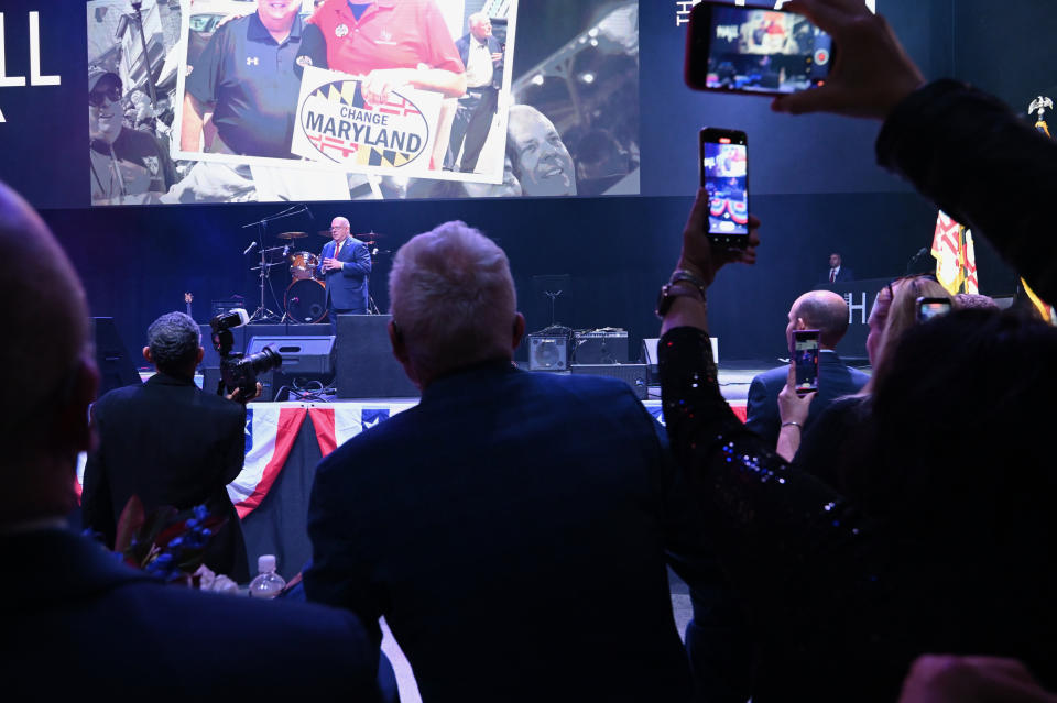 A view over the audience showing Gov. Larry Hogan in the distance in front of a large screen showing him campaigning.