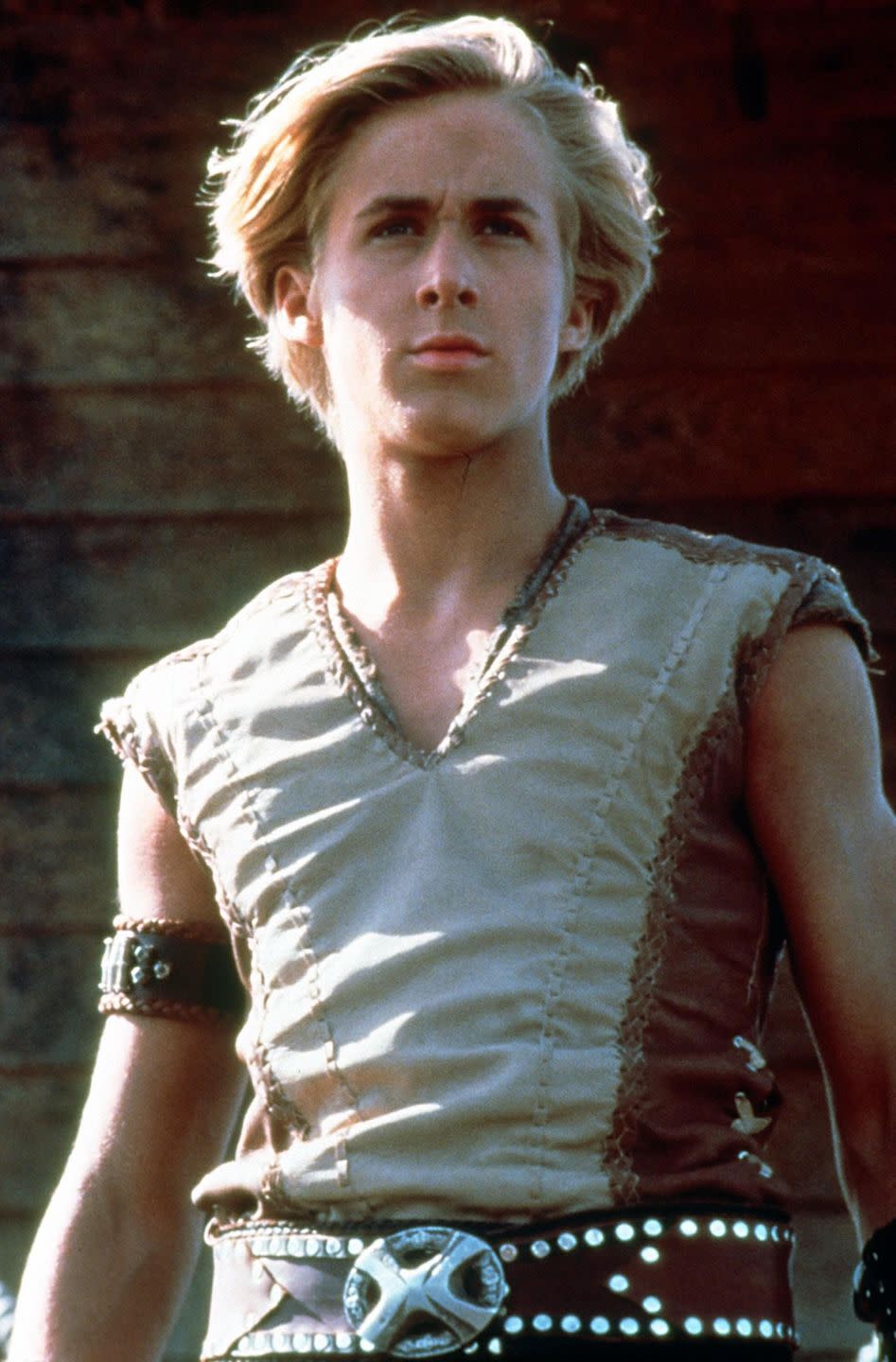 ryan gosling in character for young hercules, he wears a sleeveless tan top with an armband and a black belt