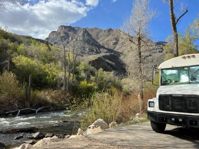 The bus parked next to a river with trees and mountains in the background