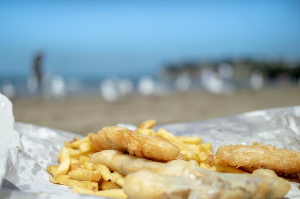 A close-up photograph of fish and chips on paper with lurking seagulls on the beach in the background