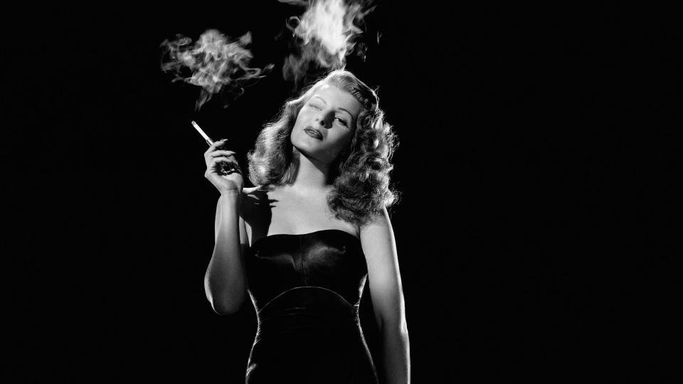 rita hayworth smokes a cigarette and stands in a dark strapless dress, she holds a fur jacket by her leg