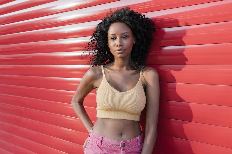 a young black woman wearing a knitted yellow crop top that exposes her stomach leaning against a red shop front shutter