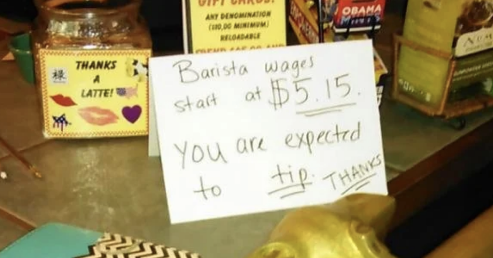 Handwritten sign states "Barista wages start at $5.15. You are expected to tip. Thanks" on a cafe counter