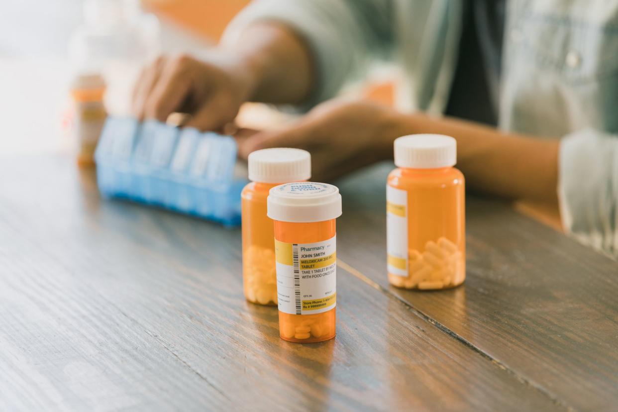 The unrecognizable man in the background of the photo fills his pill box with the medicines in the prescription bottles in foreground.