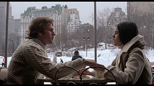 Oliver (Ryan O'Neal) and Jenny (Ali MacGraw) sit at a table drinking coffee in front of a window with a snowy cityscape outside