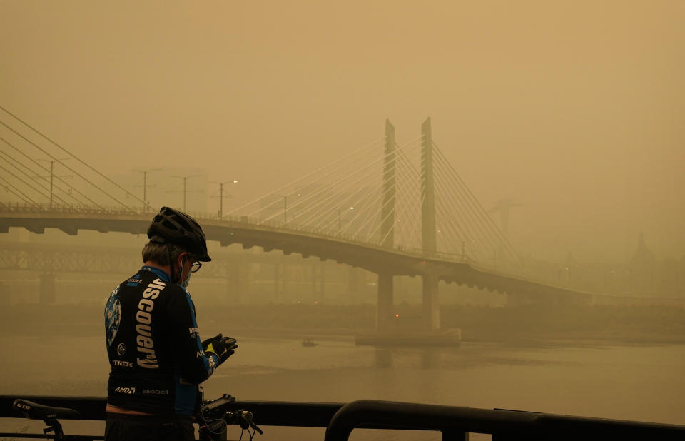 FILE - In this Sept. 12, 2020, file photo, a man stops on his bike along the Willamette River as smoke from wildfires partially obscures the Tilikum Crossing Bridge in Portland, Ore. Two unusual weather phenomena combined to create some of the most destructive wildfires the West Coast states have seen in modern times. (AP Photo/John Locher, File)