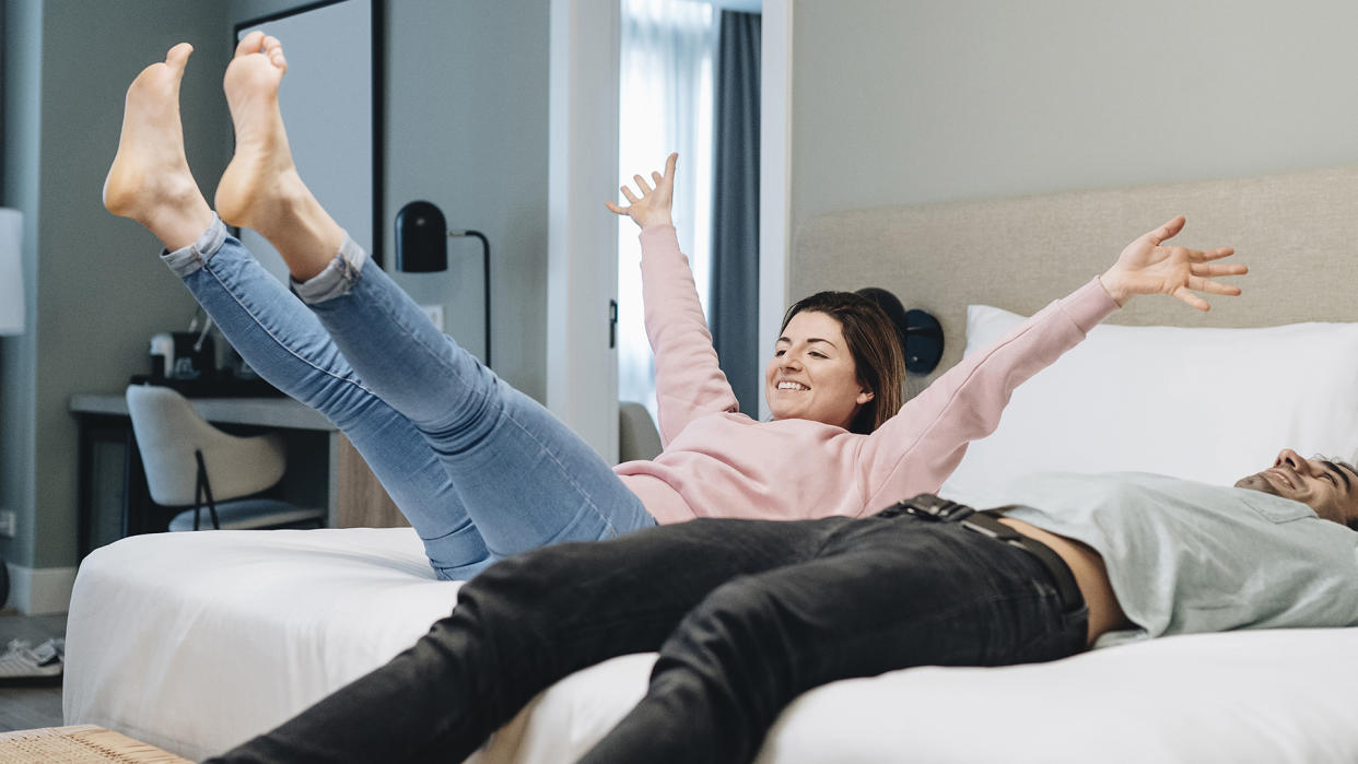  A man lies down on a bed while a woman jumps onto the bed next to him . 