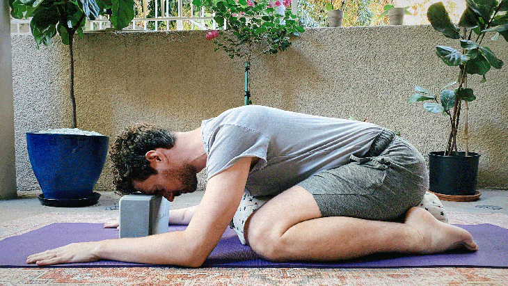 Man kneeling on his yoga mat on a bolster with a block beneath his forehead in supported Child's Pose