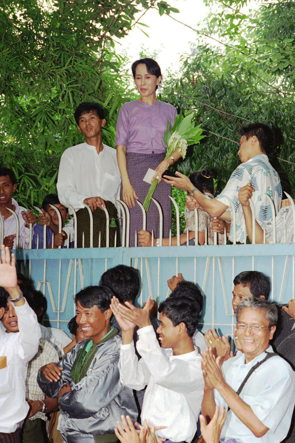 FILE - Aung San Suu Kyi, top center, stands behind her gate to greet a crowd of about 1,000 people who came to see her in Rangoon, Burma on July 11, 1995. Myanmar court on Monday, Dec. 6, 2021, sentenced ousted leader Suu Kyi to 4 years for incitement and breaking virus restrictions, then later in the day state TV announced that the country's military leader reduced the sentence by two years. (AP Photo/Stuart Isett, File)