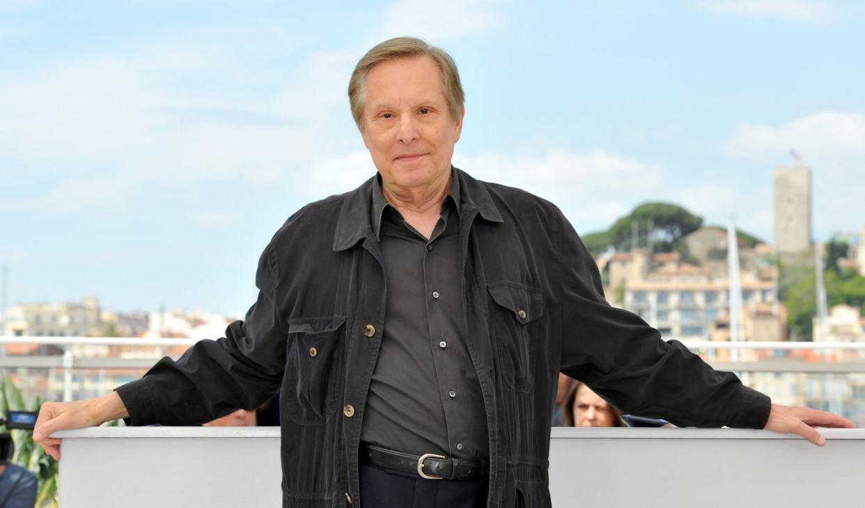 william friedkin, a mature man stands looking at the camera with a neutral facial expression, he wears a black shirt, trousers and jacket