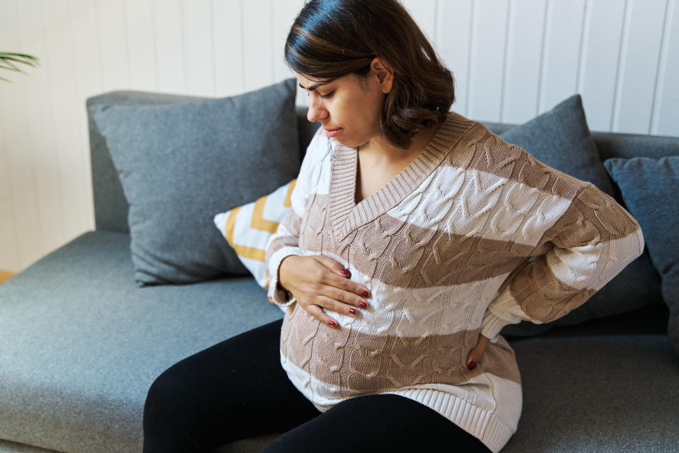 A heavily pregnant Asian woman wearing a white and brown striped jumper sits on a sofa while holding one hand against her back as though she is experiencing back pain