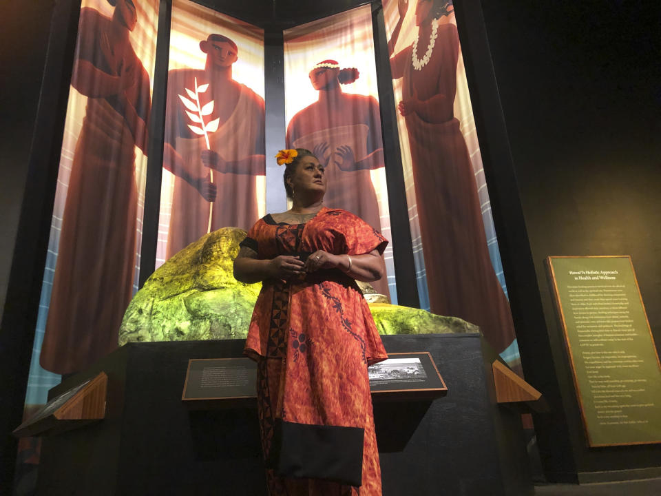 Hinaleimoana Wong-Kalu, one of the curators of the new Kapaemahu exhibit at Bishop Museum, poses for a photo in Honolulu on Thursday, June 16, 2022 in front of pictures of four healers who visited Hawaii from Tahiti more than 500 years ago. The exhibit draws attention to the stories of the healers, who were "mahu" or individuals who presented themselves as a mixture of male and female, and highlights gender fluidity's deep roots in Polynesia. (AP Photo/Audrey McAvoy)