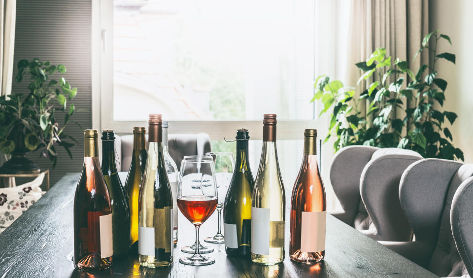 Variety of wine bottles with mock up label and glasses on table in modern living room at window. Home party preparation