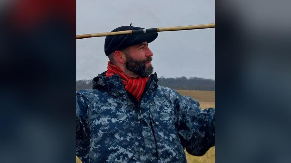 Here we see a man with a beard who is wearing a beret, orange scarf, and blue waterproof jacket. He is holding a spear, ready to throw it.