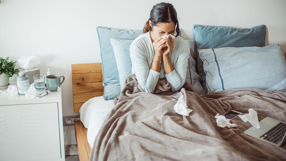 A woman with long dark hair blows her nose in bed because she is sick