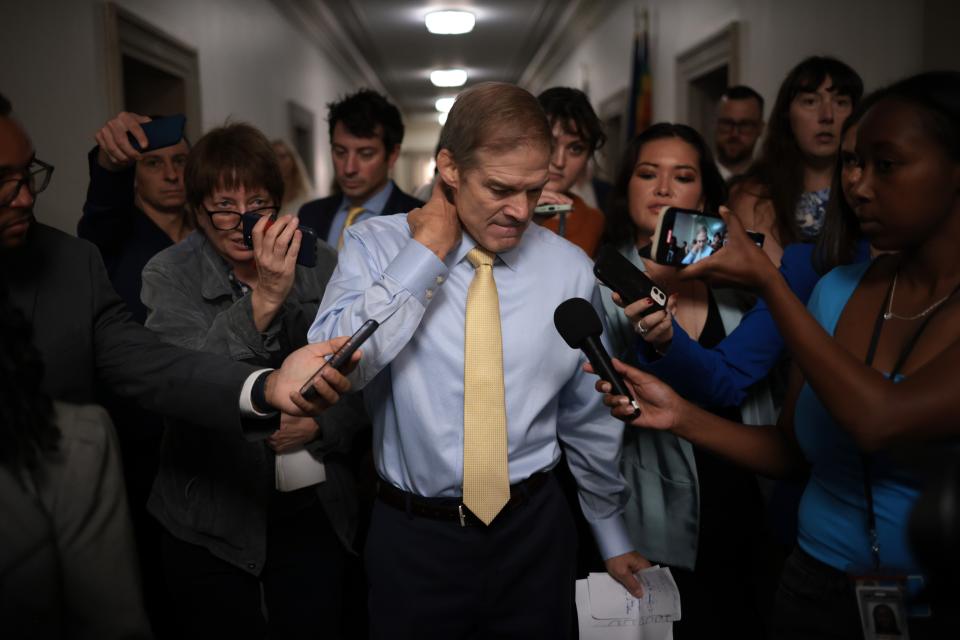 Rep. Jim Jordan, standing in a hallway surrounded by reporters holding out their microphones and voice recorders, rubs the back of his neck and looks downward.