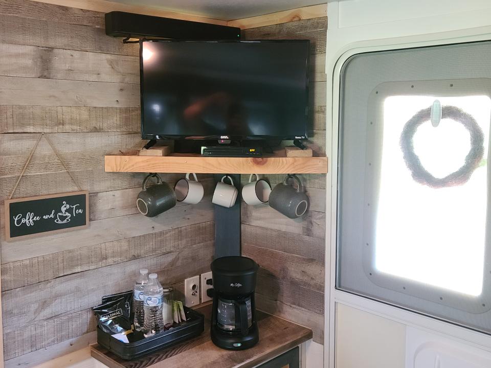 A TV on a catty-cornered shelf. There are coffee mugs hanging from the bottom of the shelf and a coffee pot and water bottles on a cabinet below. There is a door with a wreath to the right.