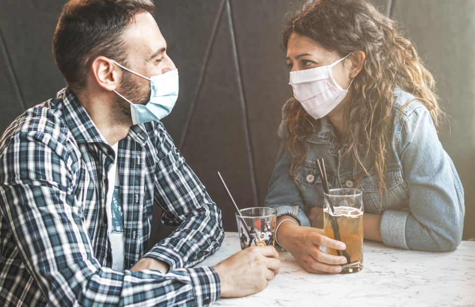 Engaged couple sitting in a caffe bar with surgical masks during the coronavirus pandemic - prevention and social distancing concept