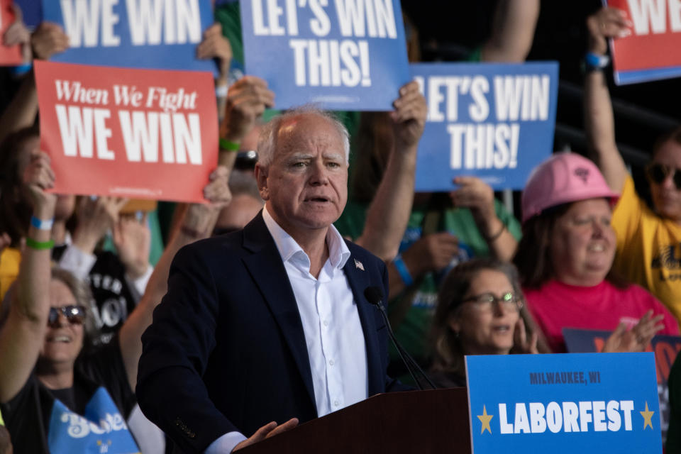 Tim Walz speaks at a podium marked "Laborfest" in Milwaukee, WI. Supporters in the background hold signs reading "LET'S WIN THIS!" and "When We Fight, We Win."