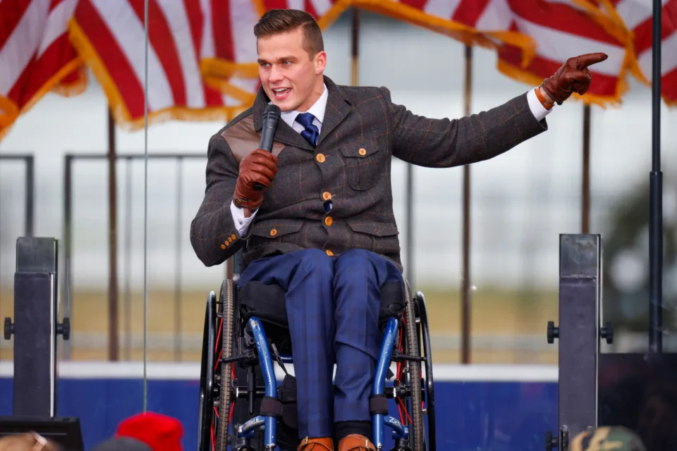 Rep. Madison Cawthorn at the microphone in his wheelchair on a podium with a backdrop of flags.