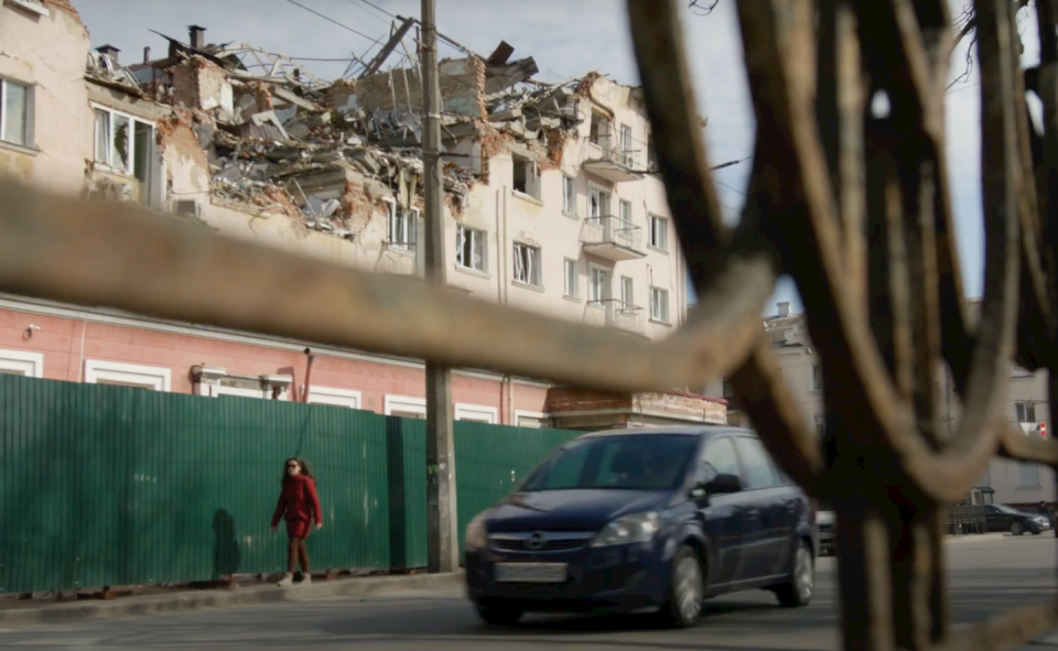 Woman in red coat walking along sidewalk as a destroyed building looms over her.