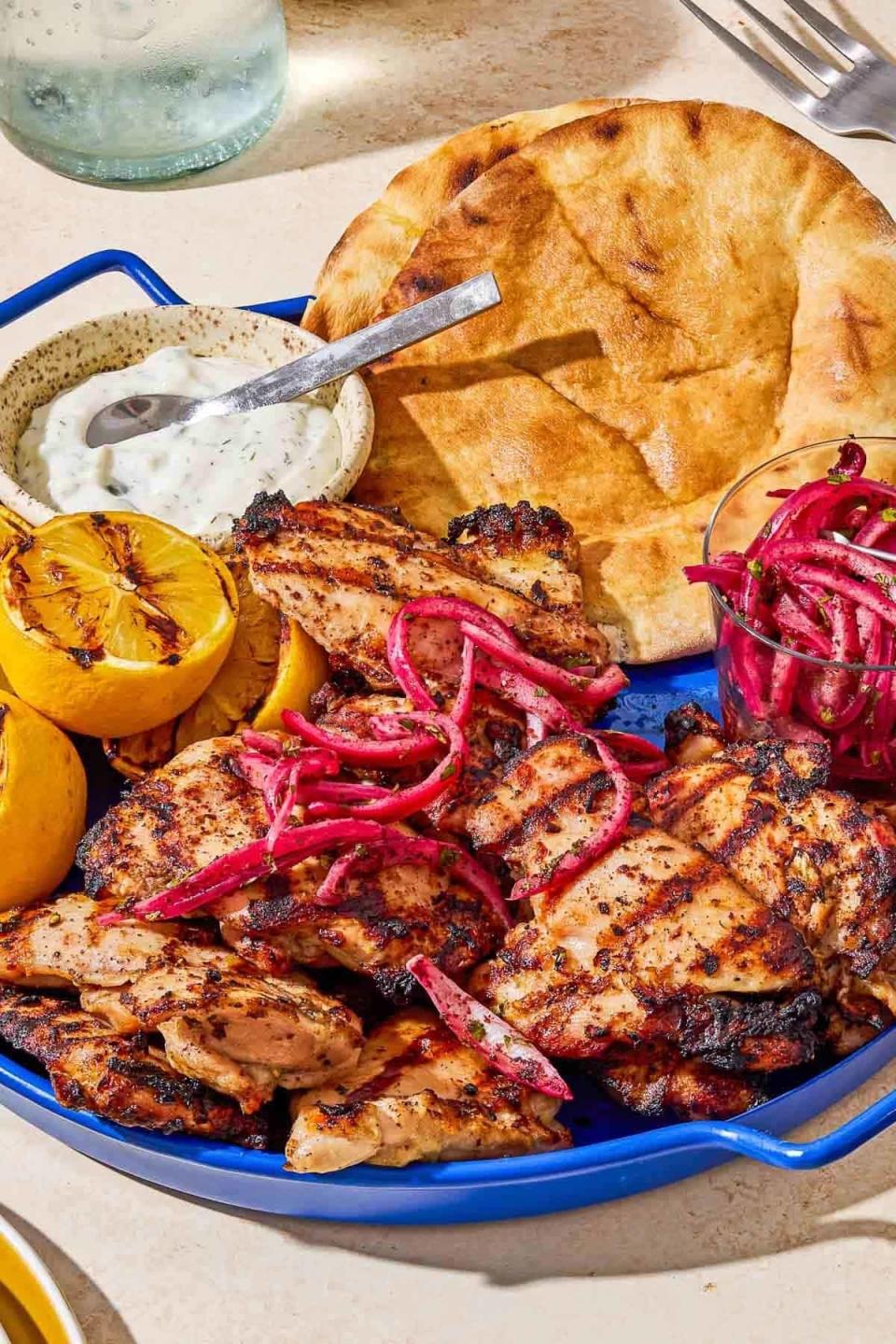 A plate with grilled chicken pieces, grilled lemons, a bowl of yogurt dip, flatbread, and pickled red onions. A fork and water glass are in the background