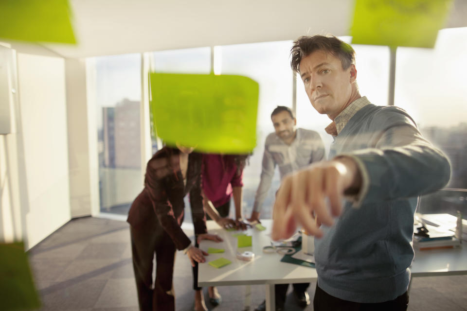 Group of diverse multi-ethnic Hispanic Black Middle Eastern Caucasian business people designers planners engineers architects consultants in contemporary modern meeting conference table room looking at post-it sticky notes on glass wall with layouts designs plans to review make plans in innovation creativity consulting business in studio work room preparation decisions choice critique feedback complexity collaboration teamwork community expertise dusk afternoon ideas brainstorming cooperation cityscape city downtown urban leadership leader