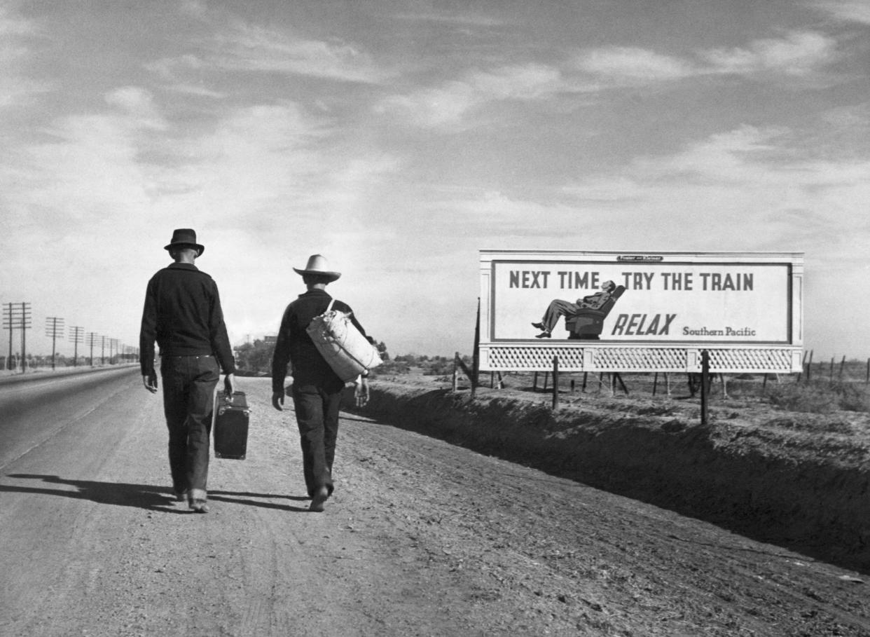 Two Dust Bowl refugees walk along a highway towards Los Angeles. passing by a billboard imploring them "Next Time Try the Train - Relax".