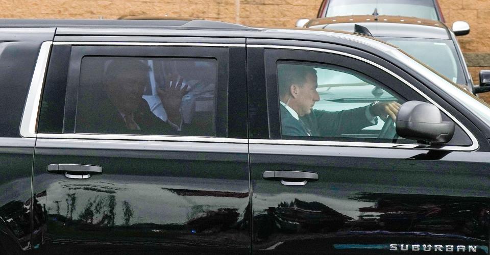 Through tinted glass of a Suburban, former President Donald Trump waves to supporters at Wheels Up Private Jet Center ahead of a fundraiser in Indian Hill later in the day