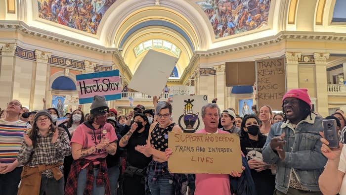 Dozens of trans people hold up signs reading "here to stay" over a handpainted trans flag, and "support and defend trans and intersex lives" and "trans rights are human rights" on pieces of cardboard at a rally at the Oklahoma state capitol