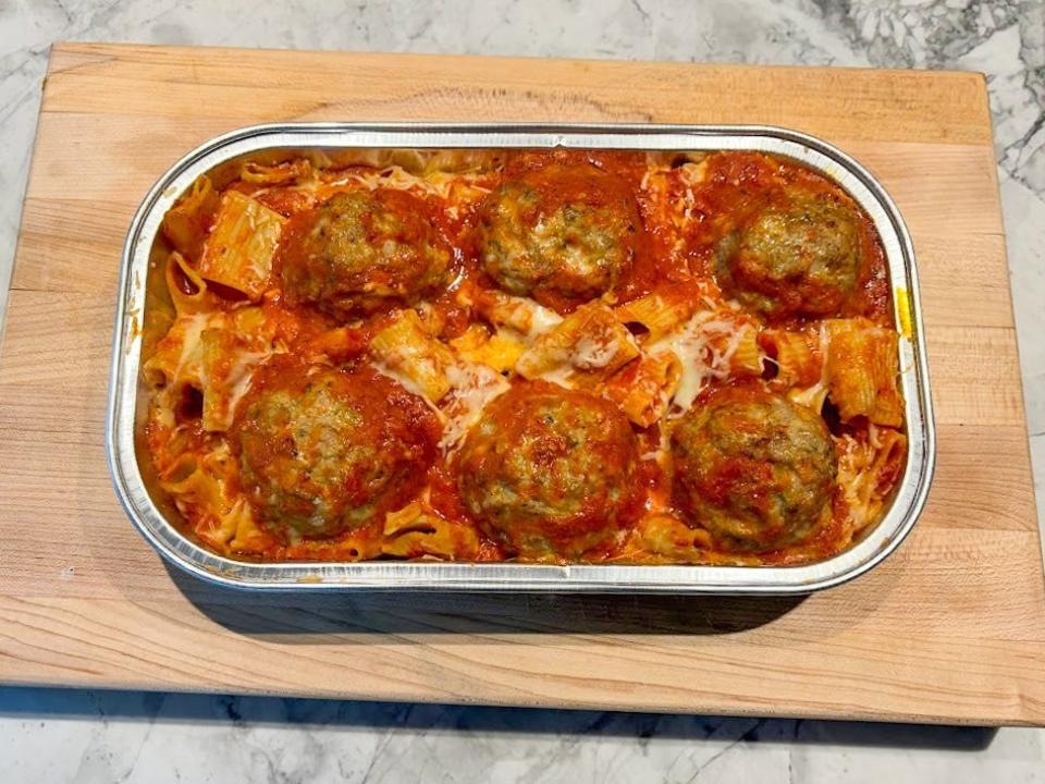 A silver tray filled with rigatoni pasta, six large meatballs, and melted cheese and bubbling red sauce set on a wooden cutting board