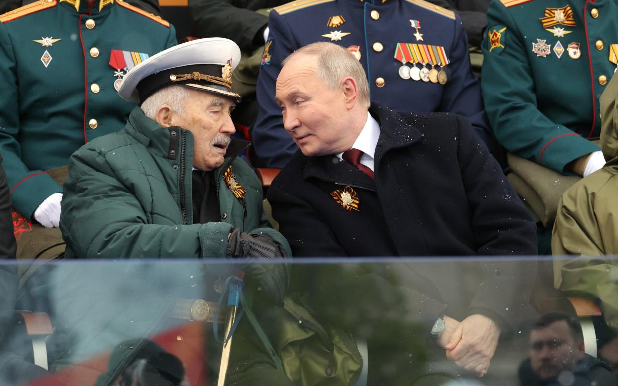 Putin smiles wryly as he speaks to an elderly serviceman seated next to him