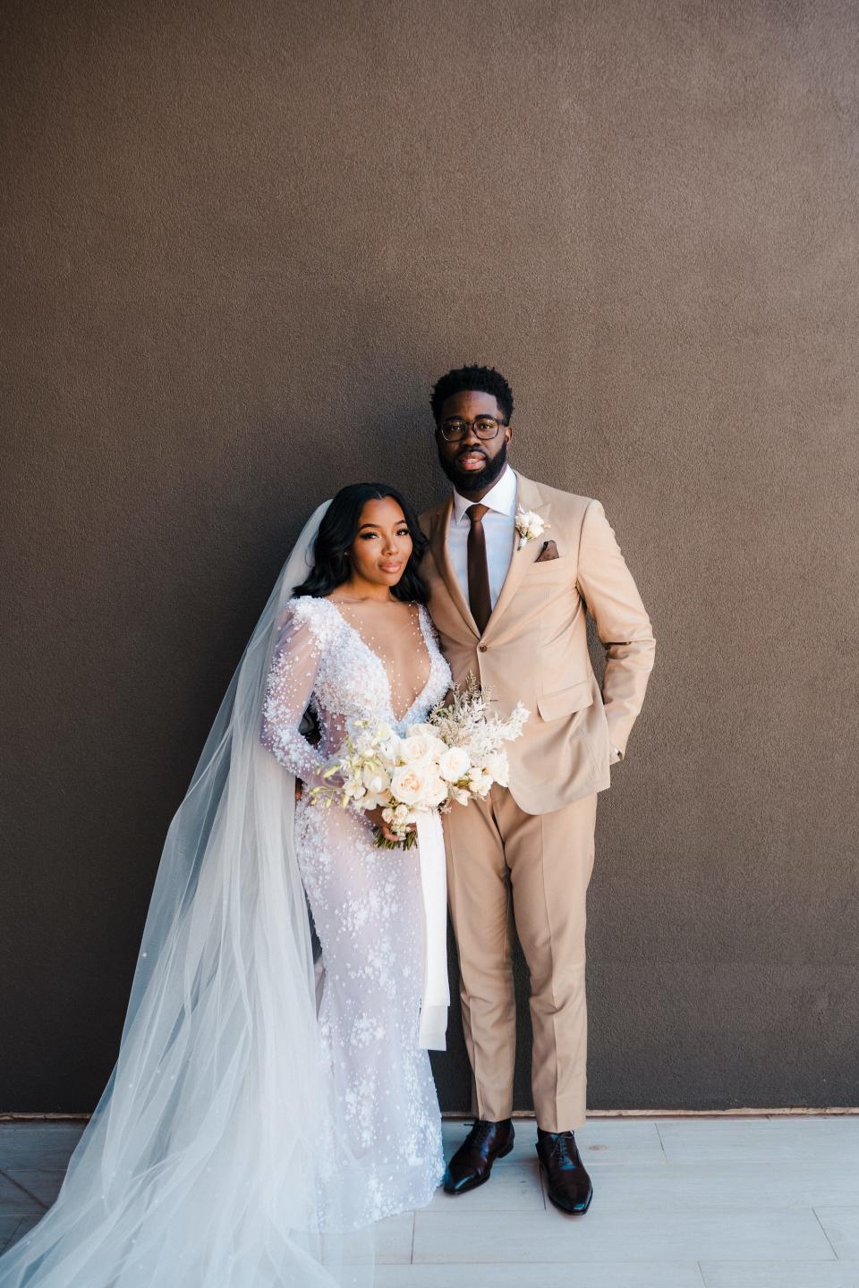 A bride and groom pose in front of a brown wall.