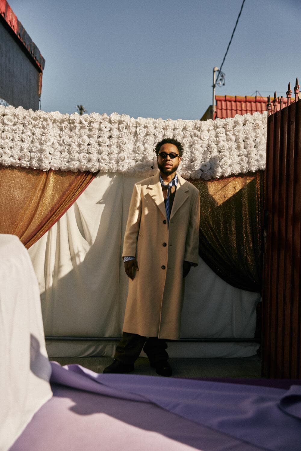 A man stands outside in a long beige coat and sunglasses.