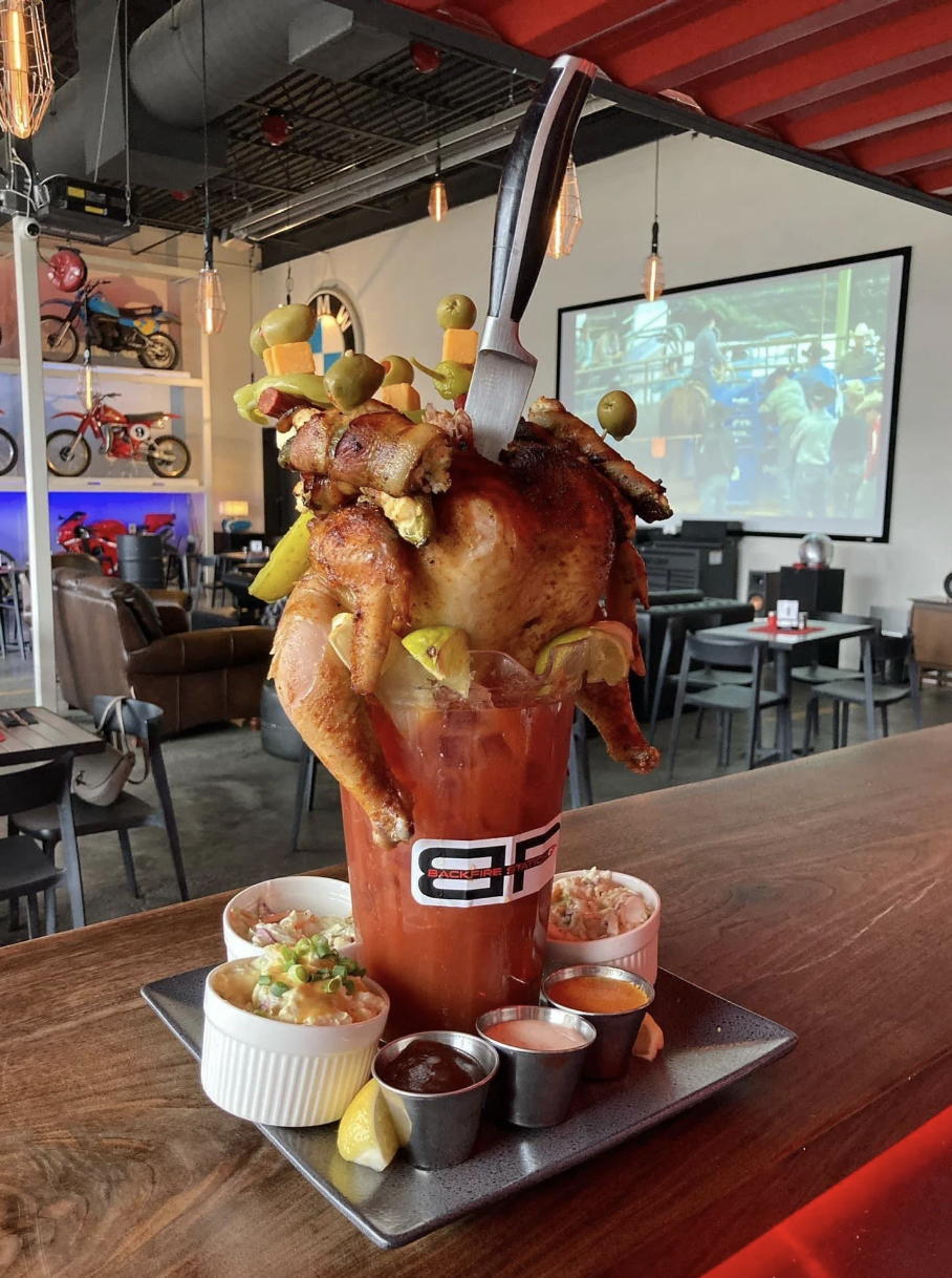 A full chicken with bacon and other condiments shoved inside a cup; there's too much food for the cup, so it's spilling out the sides