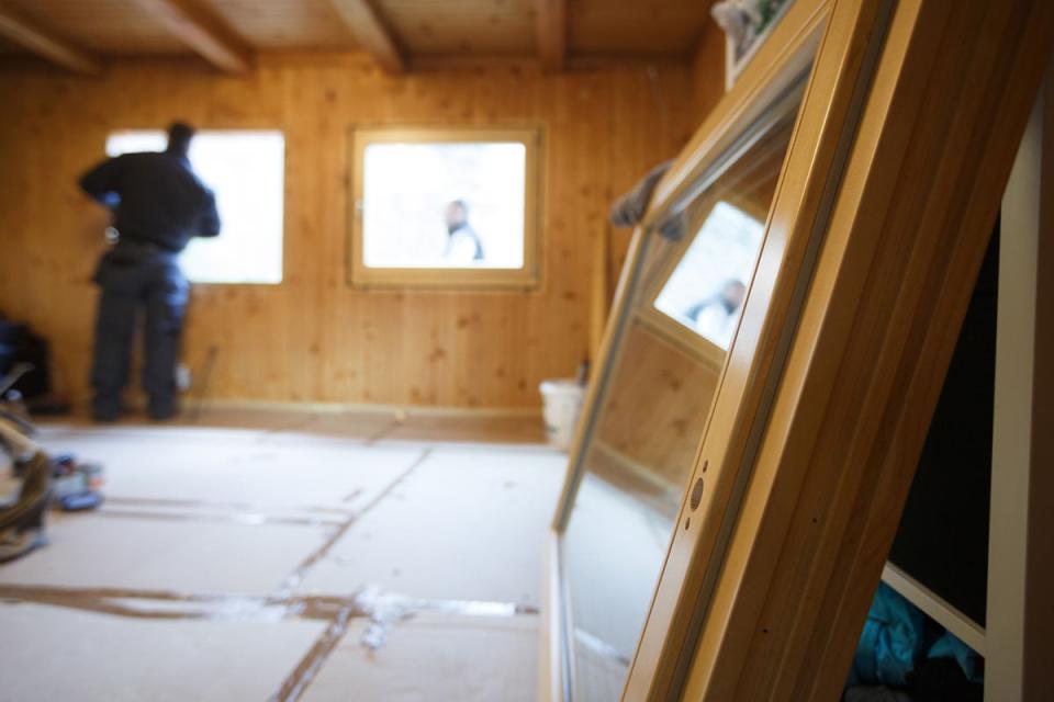 A worker in a blue jumpsuit looks out the window of a wooden interior under construction.