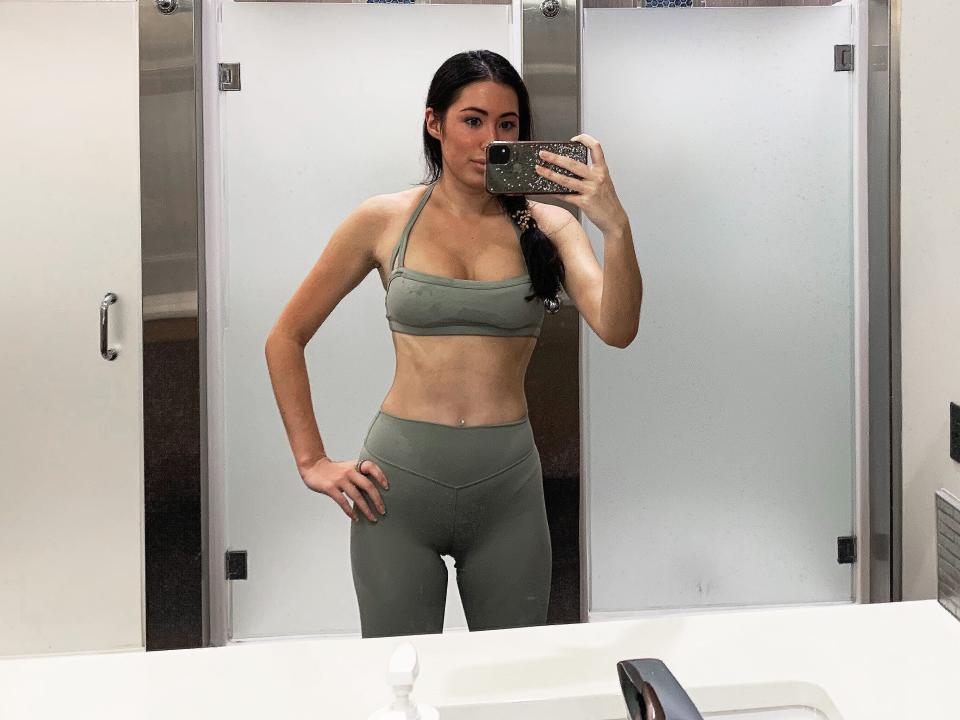 The writer wears a light-green crop top and yoga pants and stands in a bathroom mirror with her hand on her waist