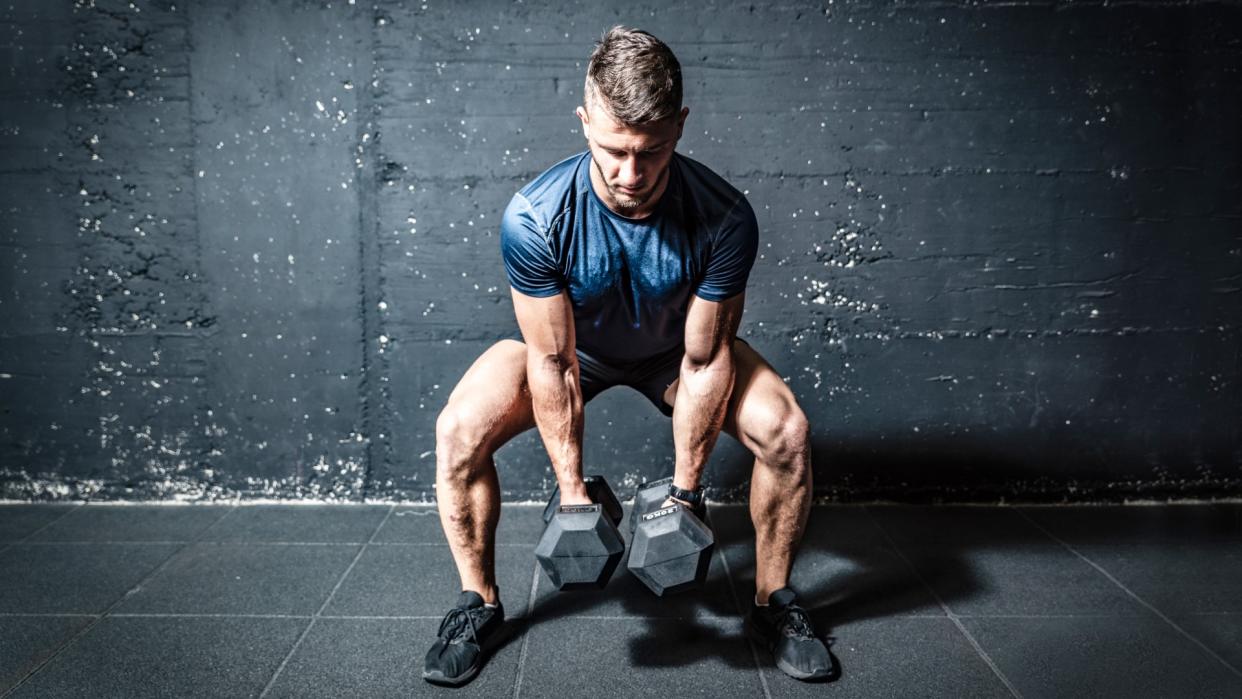  Man in squat position holding two dumbbells between legs . 