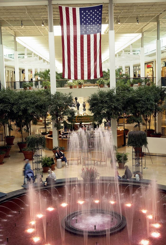 A large American flag hangs in a shopping mall atrium with a central water fountain and surrounding plants