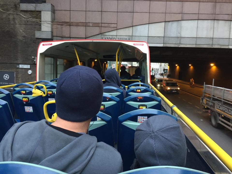 View from behind of a man in a blue sweatshirt and beanie and a child in a blue beanie on the London Hop-On Hop-Off bus, which has blue seats and no roof, going under the Cannon Bridge