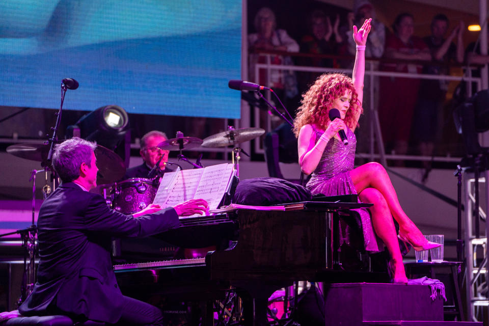 Bernadette Peters sitting on a piano with a microphone in her hand.