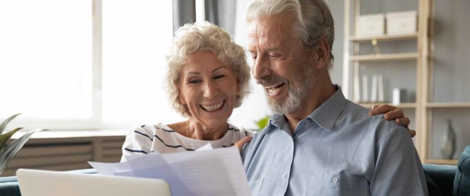 Cheerful elderly couple sit on couch, smiling and looking at bills