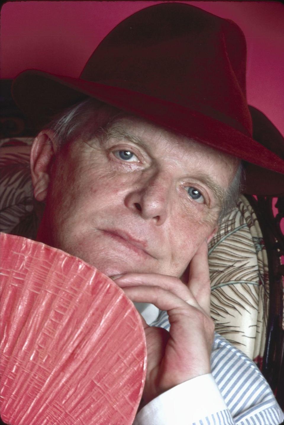 truman capote looks at the camera as he caresses his face with one hand, he wears a red hat and holds a pink fan below his face